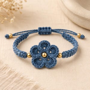 May include: A blue macrame bracelet featuring a central crocheted flower motif with a gold-toned metallic bead at its center. The band is constructed from braided blue cord, accented with two additional gold-toned beads and an adjustable sliding knot.