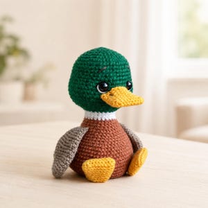 Op de afbeelding: Een gehaakt speelgoed eendje met een donkergroene kop, een witte halsring, een bruin lichaam en grijze vleugels. De eend heeft een felgele snavel en gele voeten, met grote zwarte veiligheidsogen die zorgen voor een heldere uitstraling.