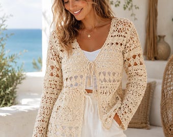 Può includere: Un cardigan a maniche lunghe in crochet color beige caratterizzato da un motivo traforato con disegni geometrici a rombi e una chiusura frontale a laccio con nappe. Il capo è indossato sopra un top bianco semplice ed è lavorato a maglia.