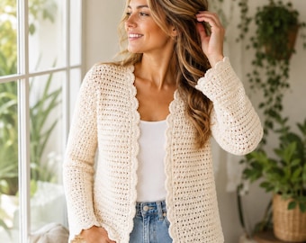 May include: A cream-coloured, long-sleeved crochet cardigan featuring a scalloped edge along the front opening and cuffs. The garment is worn over a plain white top and light blue denim jeans, showcasing a textured, open-stitch knit pattern.