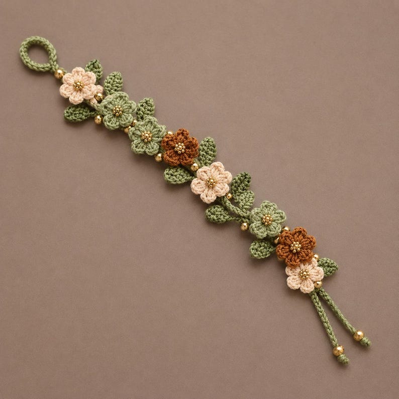 Pu&ograve; includere: Un braccialetto all'uncinetto con una serie di piccoli fiori in filato color crema, verde salvia e marrone. Il motivo floreale &egrave; impreziosito da minuscole perline metalliche dorate al centro di ogni fiore e tra le foglie, terminando con due cordini.