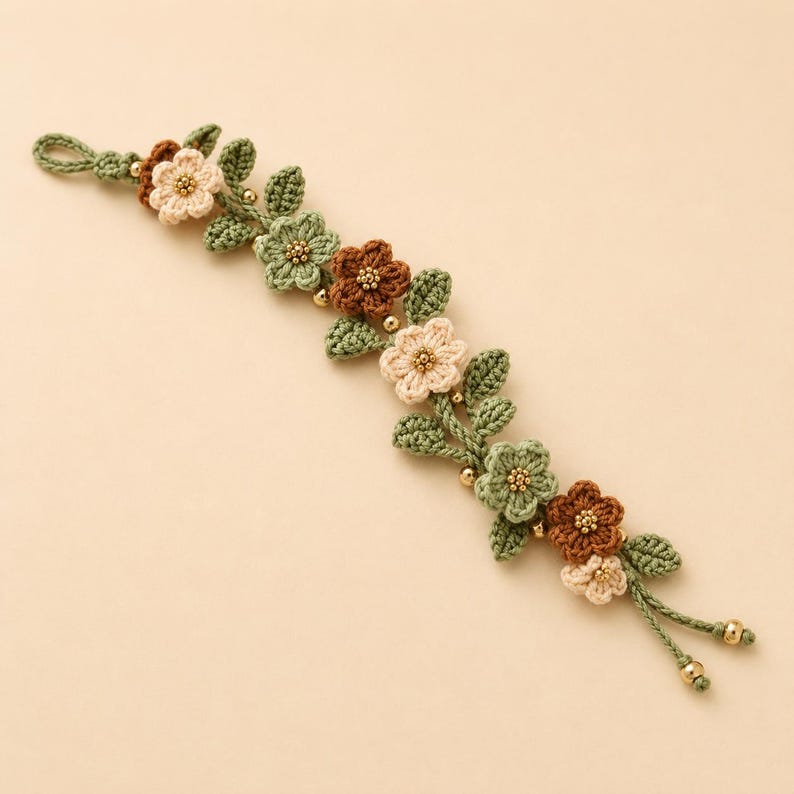 Pu&ograve; includere: Un braccialetto all'uncinetto con un design a tralcio di vite con foglie verde salvia e piccoli fiori nelle tonalit&agrave; crema, verde chiaro e marrone. I centri dei fiori sono accentuati da minuscole perline dorate e le estremit&agrave; hanno perline.
