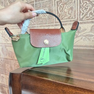 May include: A small, olive green handbag with a brown flap and handle. The bag has a gold-colored zipper pull and a small green tag that reads "LE PLIAGE GREEN". The bag is held by a person.