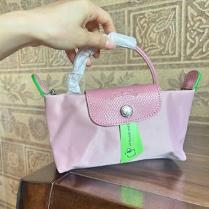 May include: A pink handbag with a leather flap and handle. The bag has a silver button closure and a green tag. The handle is wrapped in white paper. The bag is held by a hand.