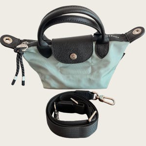 May include: A light blue handbag with black leather handles and a flap closure. The bag includes a detachable black shoulder strap. The bag has a silver-tone metal button and accents. The bag is designed for everyday use.
