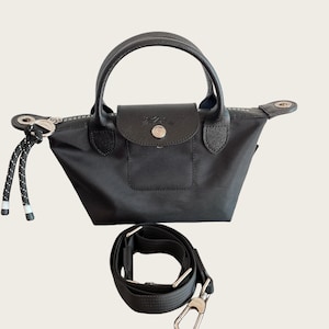 May include: Black handbag with a top handle and a detachable shoulder strap. The bag features a flap closure with a silver button and a small front pocket. The bag has a black and white rope detail on the zipper.
