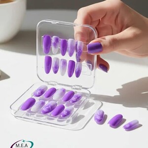 Peut inclure: Une boîte en plastique transparente contenant un ensemble de faux ongles violets et blancs ombrés. Les ongles sont de forme ovale et ont une finition brillante. La boîte est ouverte et quelques ongles sont éparpillés. Le logo "M.E.A STUDIO AURA" est visible.