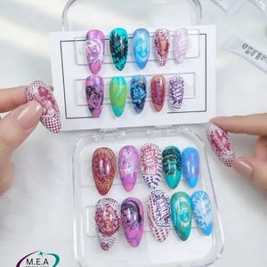Peut inclure: Une boîte en plastique transparente contient un ensemble d'ongles artificiels décorés et colorés. Chaque ongle présente un design unique, comprenant des personnages de dessins animés et des motifs abstraits. Les ongles sont de différentes couleurs, comme le rose, le bleu et le vert. Le logo "M.E.A STUDIO AURA" est visible.