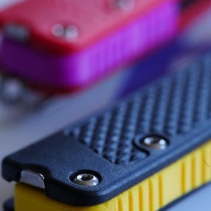 Könnte beinhalten: Zwei Multifunktionswerkzeuge mit strukturierten Griffen. Das vordere Werkzeug hat eine schwarze Deckplatte mit gelbem, geripptem Seitenteil und silbernen Schrauben. Das hintere Werkzeug hat eine rote Deckplatte mit violettem Seitenteil.