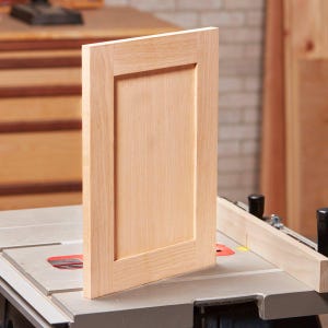 Könnte beinhalten: Eine unfertige, hellfarbene Holztür für einen Schrank mit einer eingelassenen Platte. Die rechteckige Tür hat einen erhabenen Rahmen und steht auf einer Tischkreissäge. Die Holzmaserung ist sichtbar.