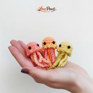 Könnte beinhalten: Drei handgefertigte Häkelquallen in Rosa, Orange und Gelb. Jede Qualle hat schwarze Knopfaugen und lange, fließende Tentakel. Das Loop Paws-Logo befindet sich oben rechts.