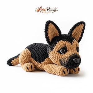 Puede incluir: Un perro pastor alemán tejido a ganchillo en posición de descanso. El perro es principalmente marrón y negro, con una nariz negra y ojos grandes y expresivos. El logotipo de Loop Paws está en la esquina superior izquierda.