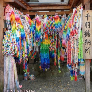 以下が含まれることがあります： 木製の構造から吊り下げられた、カラフルな折り紙の鶴の展示。鶴は様々な色で、虹色の効果を生み出しています。日本語の文字が書かれた看板が見えます。画像には「KATANISIM - MADE IN SILENCE」というテキストが含まれています。
