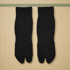 以下が含まれることがあります： 黒色の足袋ソックス一組。つま先が分かれたデザインです。リブ編みのニット素材でできており、ニュートラルな背景に展示されています。伝統的な日本の履物と一緒に着用することを目的としています。