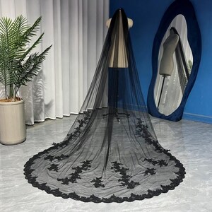 May include: A black bridal veil with floral lace edging, displayed on a mannequin. The veil's sheer fabric is visible, with the lace detail adding a decorative element. The veil is long, with the lace detailing along the bottom edge.
