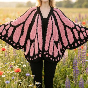 Op de afbeelding: Een roze en zwarte cardigan in de vorm van een vlindervleugel. De open trui heeft een V-hals en is gemaakt van een zachte, getextureerde brei. De vleugels zijn roze met zwarte accenten en een gestippelde rand. De cardigan wordt gedragen over een zwarte top en legging.