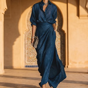 May include: A dark blue, two-piece outfit consisting of a button-down shirt and a wrap skirt. The shirt has long sleeves and a collared neckline. The skirt has a high waist and a draped design. A small, brown clutch is held in the hand.