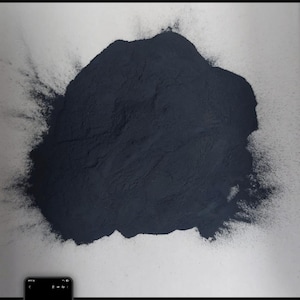 Puede incluir: Un polvo azul oscuro, posiblemente un pigmento o tinte, se extiende sobre una superficie blanca. El polvo tiene una forma grande e irregular, con una textura fina. La imagen está bien iluminada, mostrando los detalles del polvo.