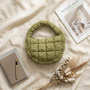May include: Olive green quilted handbag with a rounded shape and a top handle. The bag is placed on a cream-colored surface, alongside a notebook, pen, magazine, and dried flowers. The magazine reads "MODERN MUSE".