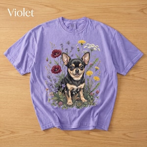 May include: Violet-colored t-shirt featuring a detailed illustration of a Chihuahua dog surrounded by wildflowers. The dog is black and tan, with large ears and a friendly expression. The flowers include red poppies, yellow dandelions, and white blooms.