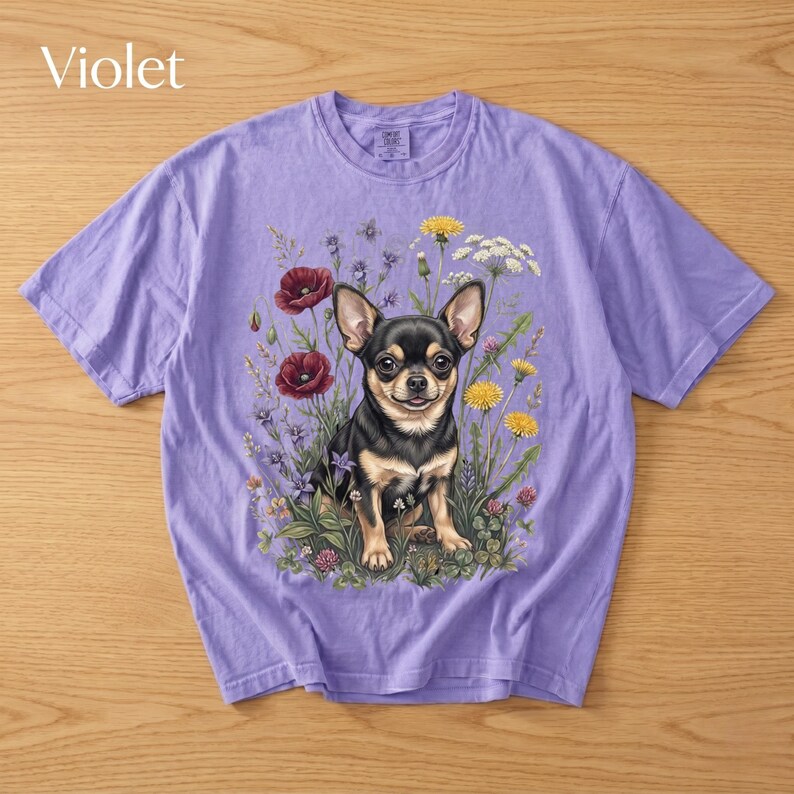 May include: Violet-colored t-shirt featuring a detailed illustration of a Chihuahua dog surrounded by wildflowers. The dog is black and tan, with large ears and a friendly expression. The flowers include red poppies, yellow dandelions, and white blooms.