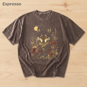 May include: Espresso-coloured t-shirt with a wolf illustration surrounded by flowers and a full moon. The wolf has golden eyes and detailed fur. The shirt is a short-sleeved crew neck.