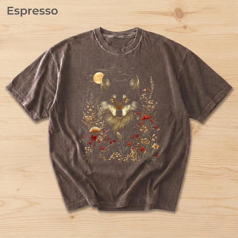 May include: Espresso-coloured t-shirt with a wolf illustration surrounded by flowers and a full moon. The wolf has golden eyes and detailed fur. The shirt is a short-sleeved crew neck.