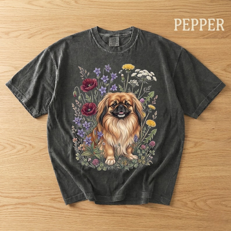 May include: Dark gray t-shirt featuring a detailed illustration of a Pekingese dog surrounded by colorful wildflowers. The dog has brown fur and is smiling. The word "PEPPER" is printed in white on the upper right.