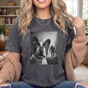 May include: Dark grey t-shirt featuring a black and white graphic of a surprised French Bulldog with a Bigfoot figure in the background. The dog has large, wide eyes. The shirt is a comfortable, casual style.