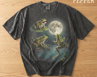 May include: Dark grey t-shirt with a whimsical illustration of three green frogs in a moonlit pond. One frog sits on a lily pad, another on the water's edge, and the third is mid-leap. The word "PEPPER" is printed at the top.