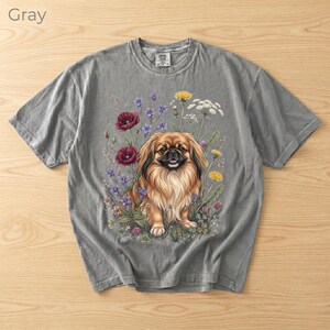 May include: Gray short-sleeve t-shirt featuring a detailed illustration of a Pekingese dog surrounded by colorful flowers. The dog has long, flowing fur and is set against a backdrop of red poppies, purple and yellow wildflowers.