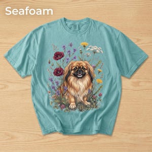May include: Seafoam-colored t-shirt featuring a detailed illustration of a Pekingese dog surrounded by colorful wildflowers. The dog has long, flowing fur and is positioned in the center of the design. The word "Seafoam" is printed at the top.