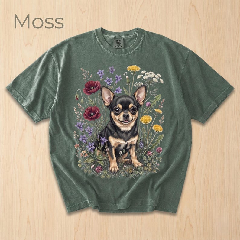 May include: Moss green t-shirt featuring a detailed illustration of a Chihuahua dog surrounded by colorful wildflowers. The dog has black and tan fur. The word "Moss" is printed in the upper left corner. The shirt has short sleeves and a crew neck.