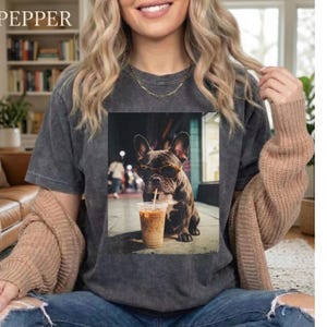 May include: Dark gray t-shirt featuring a graphic of a French Bulldog wearing sunglasses and drinking iced coffee. The word "PEPPER" is printed in the upper left corner. The shirt is worn by a person.