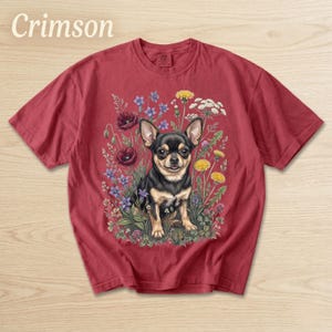 May include: Crimson-colored t-shirt featuring a detailed illustration of a black and tan Chihuahua dog surrounded by colorful wildflowers. The text "Crimson" is displayed at the top left. The shirt is a comfortable, casual style.