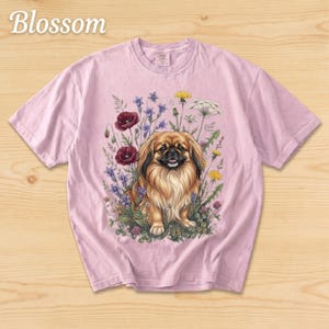 May include: A light pink t-shirt featuring a detailed illustration of a Pekingese dog surrounded by wildflowers. The dog has brown and tan fur. The word "Blossom" is printed in a script font at the top left.