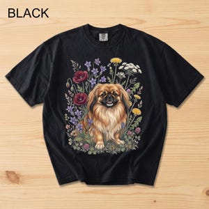May include: Black t-shirt featuring a detailed illustration of a Pekingese dog surrounded by colorful wildflowers. The dog has long, flowing brown fur and is smiling. The word "BLACK" is printed at the top left.
