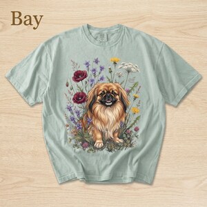 May include: A light green t-shirt featuring a detailed illustration of a Pekingese dog surrounded by colorful wildflowers. The word "Bay" is printed in the upper left corner. The dog has brown fur and is sitting in a field of flowers.