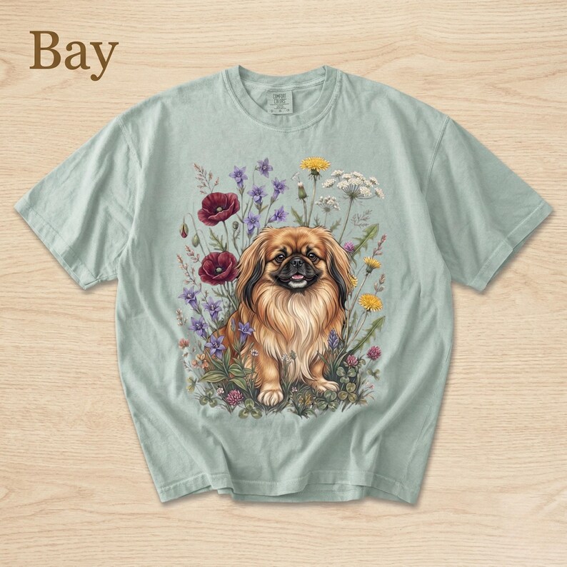 May include: A light green t-shirt featuring a detailed illustration of a Pekingese dog surrounded by colorful wildflowers. The word "Bay" is printed in the upper left corner. The dog has brown fur and is sitting in a field of flowers.