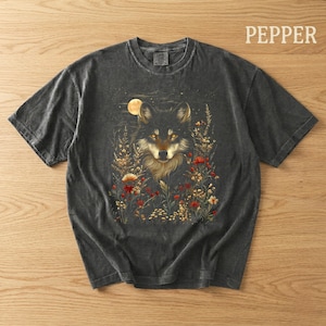 May include: Dark grey t-shirt with a detailed illustration of a wolf surrounded by flowers under a full moon. The wolf has golden eyes and a fluffy mane. The word "PEPPER" is printed in white on the upper right.