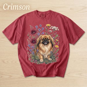 May include: Crimson-colored t-shirt featuring a detailed illustration of a Pekingese dog surrounded by colorful wildflowers. The dog has long, flowing fur and is positioned in the center of the design. The word "Crimson" is printed at the top.