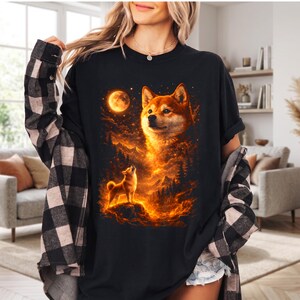 May include: Black t-shirt featuring a Shiba Inu dog design. The graphic depicts a Shiba Inu under a full moon, with fiery orange and yellow clouds and mountains in the background. The design includes a smaller Shiba Inu howling.