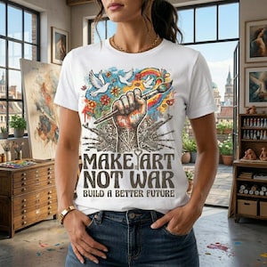 May include: White t-shirt with the text "MAKE ART NOT WAR" and "BUILD A BETTER FUTURE". The shirt features a graphic of a fist holding a paintbrush, doves, flowers, and a rainbow. The shirt is paired with blue jeans.