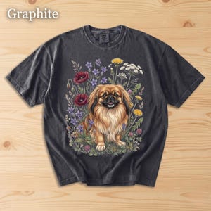 May include: Graphite-colored t-shirt featuring a detailed illustration of a Pekingese dog surrounded by colorful wildflowers. The dog has long, flowing fur and a happy expression. The flowers include red poppies, purple blooms, and yellow dandelions.
