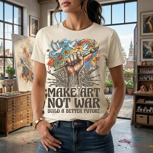 May include: Cream-colored t-shirt with the text "MAKE ART NOT WAR" and "BUILD A BETTER FUTURE". The graphic features a raised fist holding a paintbrush, doves, flowers, and a rainbow. The design is set against a backdrop of guns and barbed wire.