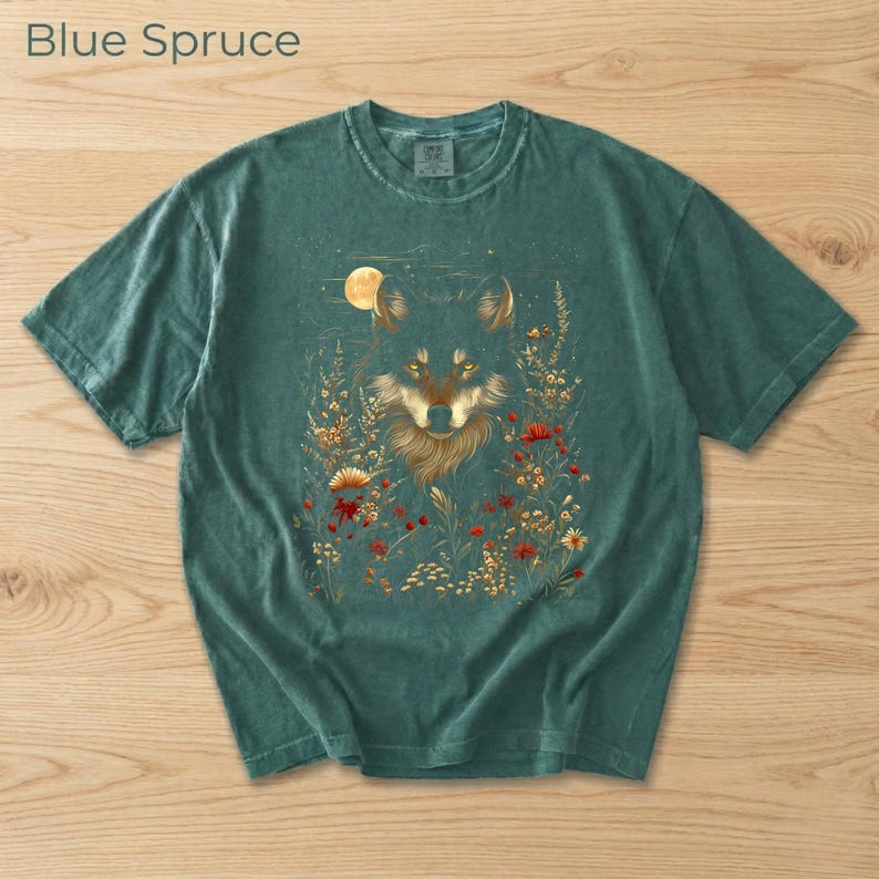 May include: Teal Blue Spruce t-shirt with a wolf design. The illustration shows a wolf with golden fur, wildflowers, and a full moon. The flowers are red, yellow, and white. The shirt's color is a muted teal, and the text "Blue Spruce" is at the top.