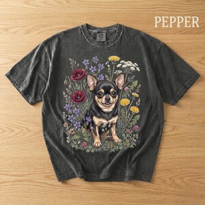 May include: Dark gray t-shirt featuring a black and tan Chihuahua dog surrounded by colorful flowers. The word "PEPPER" is printed in white on the upper right. The shirt has a vintage, washed look.