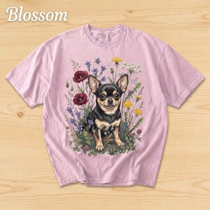 May include: A light pink t-shirt featuring a detailed illustration of a black and tan Chihuahua surrounded by wildflowers. The word "Blossom" is printed in a script font at the top left. The shirt is made of a soft material.
