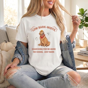 May include: White t-shirt with a graphic of a dog wearing a hat and bandana. The text reads "LOW BARK-WIDTH. SEARCHING FOR WI-BARK NO SIGNAL JUST BARK". The shirt is worn by a person.