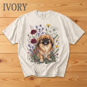 May include: Ivory-colored t-shirt featuring a detailed illustration of a Pekingese dog amidst a vibrant floral arrangement. The dog has brown and tan fur, and the flowers include red poppies, purple blooms, and yellow dandelions. The word "IVORY" is printed at the top.